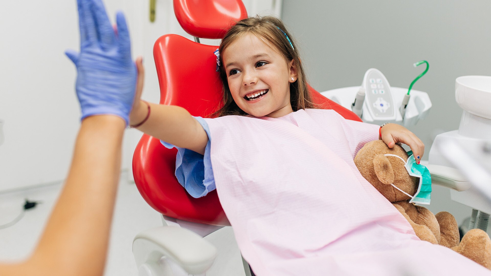 Image of a little girl who is smiling and sitting in a reddental chair, giving a high five to the doctor