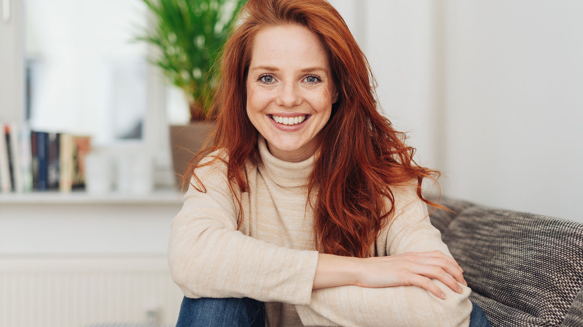 Young woman with red hair wearing a beige turtleneck sweater and smiling directly at the camera 