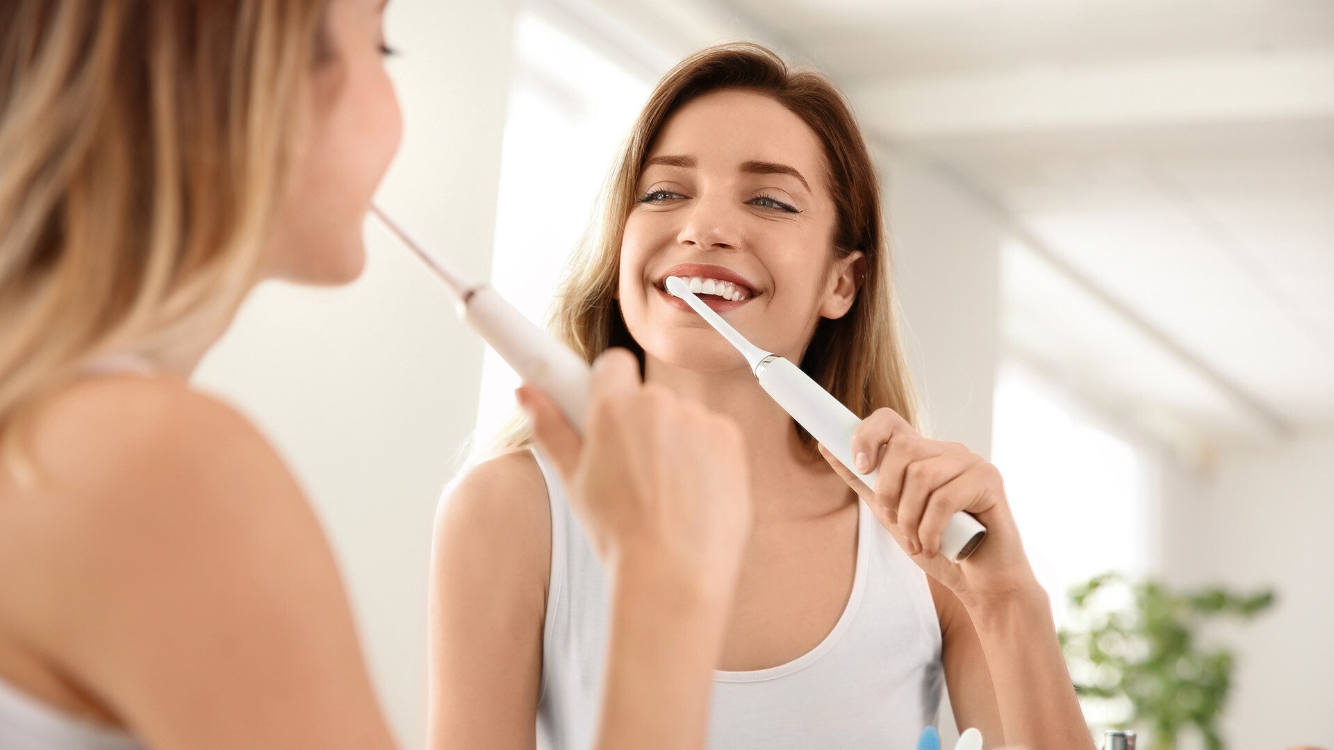 Young brunette woman in a white shirt standing in front of a mirror and brushing her teeth with an electric toothbrush 