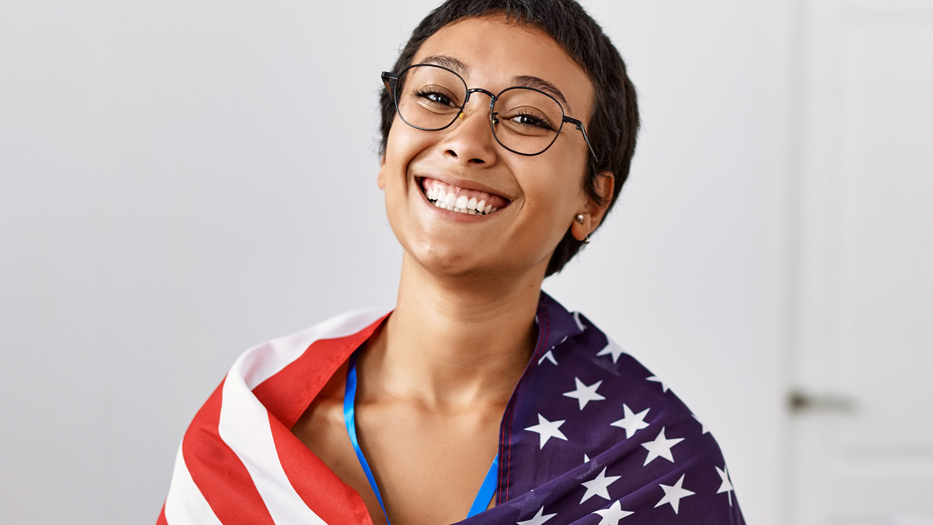 Image of a hispanic woman with short hair who is smiling with very white teeth and wearing an American flag over her shoulders