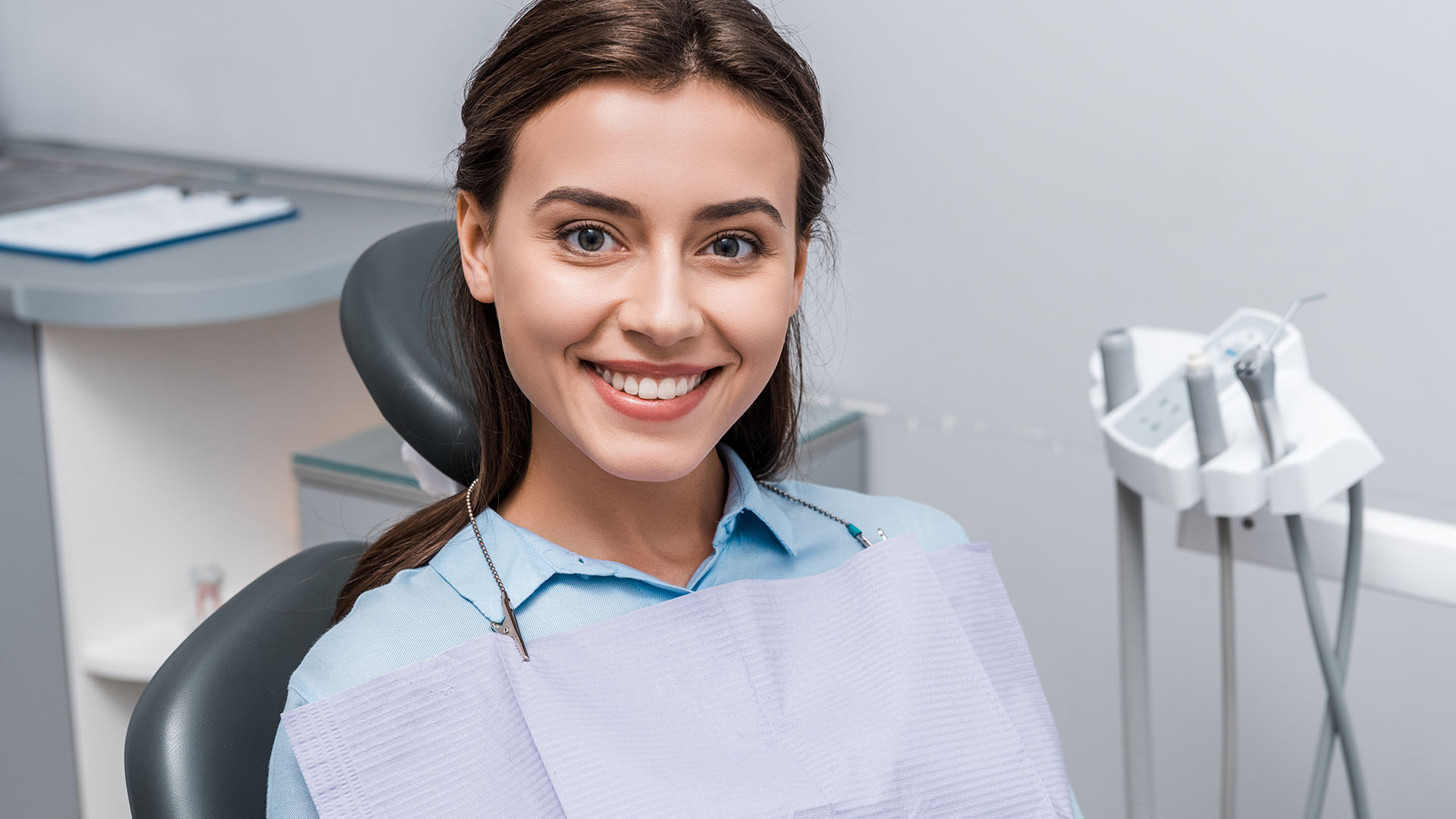 Beautiful young woman with brown hair and blue shirt sitting in a dental chair and smiling