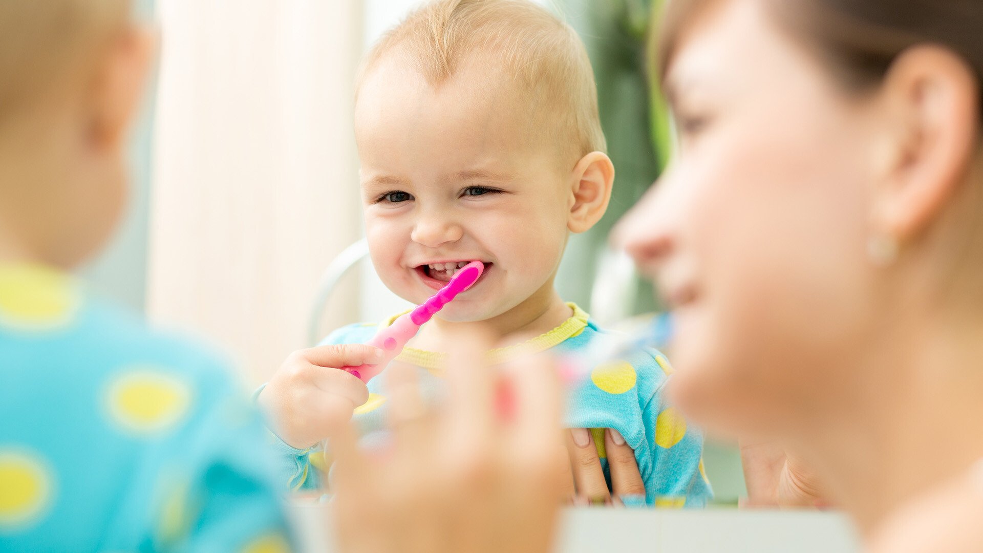 An image of a mother and child looking in a mirror and smiling while brushing the childs teeth 